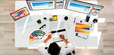 A graphic designer working on a creative project at a modern workspace, surrounded by color swatches, design tools, and dual monitors displaying website layouts.