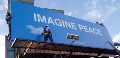 Workers installing a large blue billboard that reads 'IMAGINE PEACE,' symbolizing collaboration and professional craftsmanship