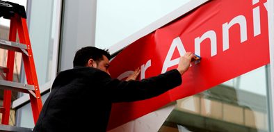 A worker installing a large red vinyl sign on a storefront window using precision tools and a ladder.