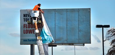 A worker performing maintenance on a large outdoor billboard using a bucket lift, removing or repairing the signage.