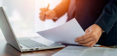 A business professional reviewing documents and taking notes at a desk with a laptop, representing paperwork review or permit application processes.