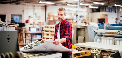 A professional worker in a print production facility examining large printed sheets, surrounded by printing equipment and machinery.